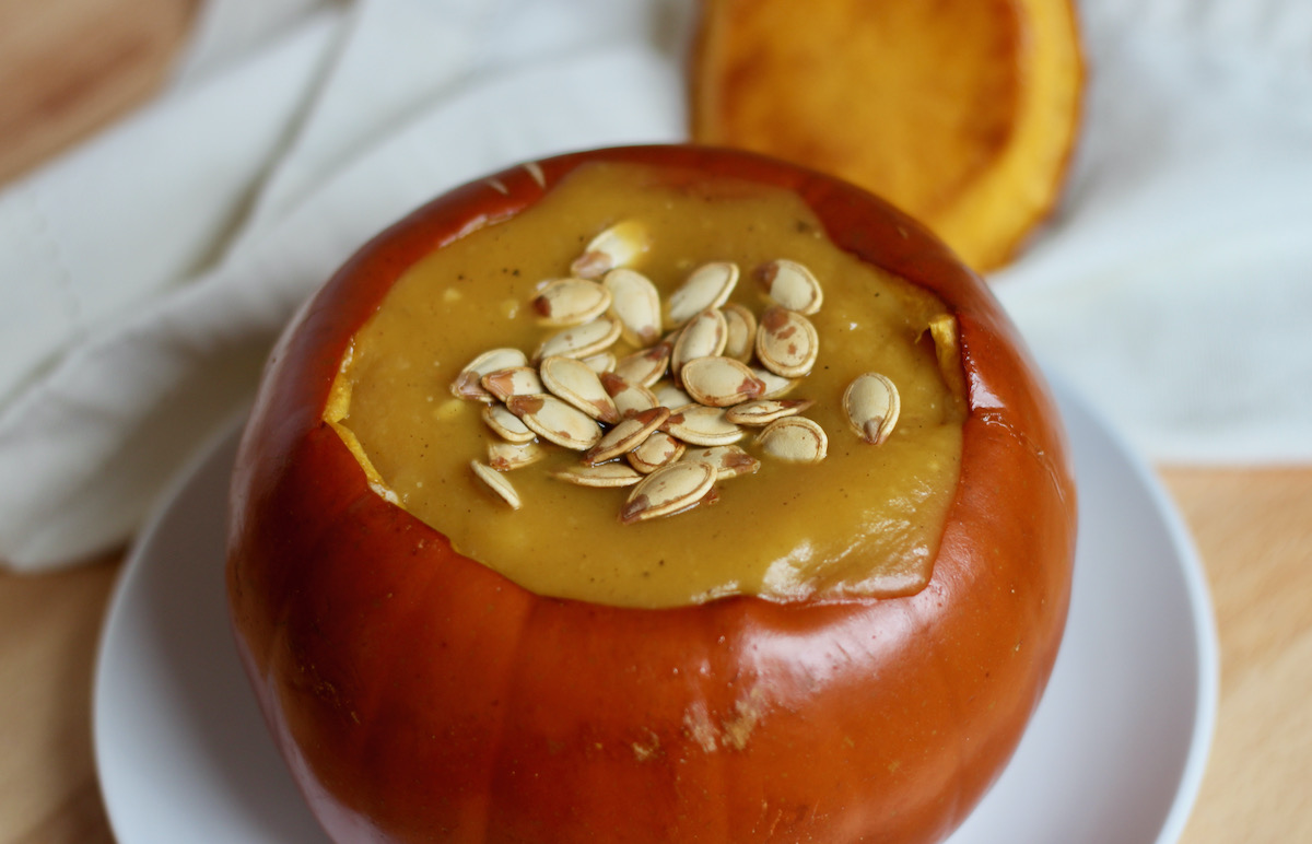 A roasted pumpkin bowl filled with pumpkin soup. The soup is topped with roasted pumpkin seeds. You can see the top of the pumpkin bowl out of focus in the background.