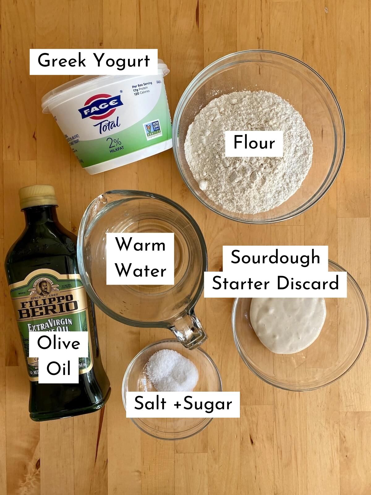 The ingredients to make sourdough discard naan bread on a wooden countertop. The ingredients are labeled with text over each one. They include flour, Greek yogurt, warm water, sourdough discard starter, olive oil, salt, and sugar.
