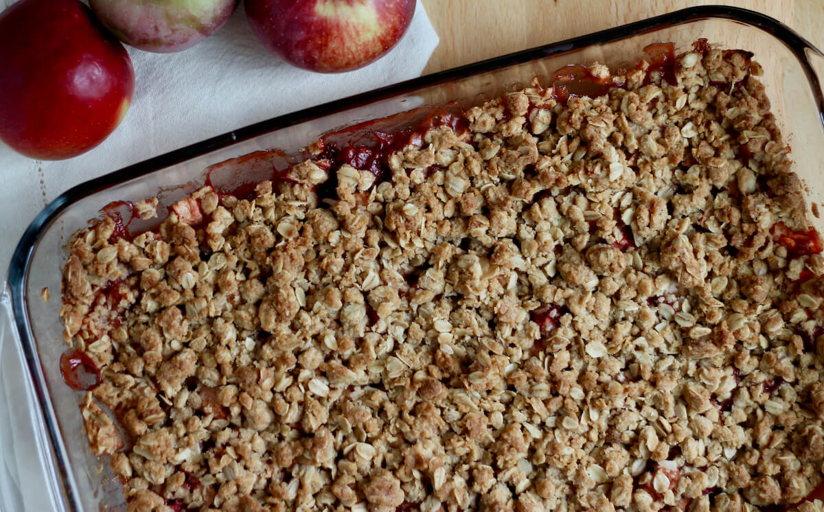 A baking dish with baked strawberry apple crisp. There are three apples out of focus next to the baking dish.