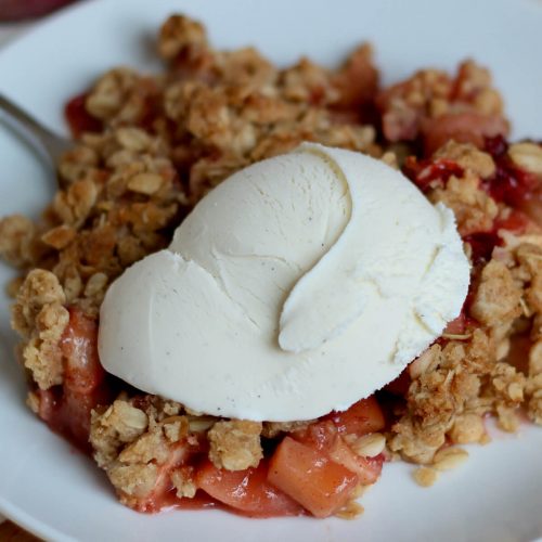 Strawberry apple crisp topped with vanilla ice cream on a small white plate. There is a fork sticking out of the crisp out of focus in the background.