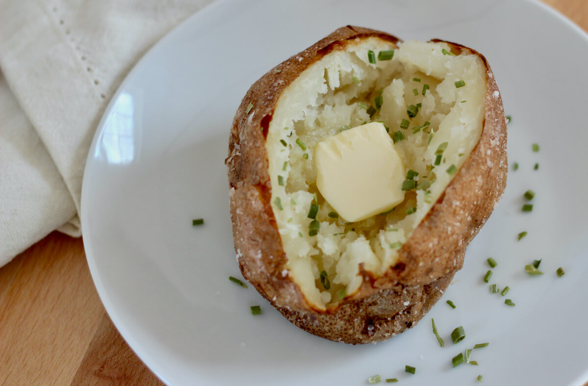 A baked potato cut in half on a small white plate. The potato is seasoned with chives and a pat of butter.