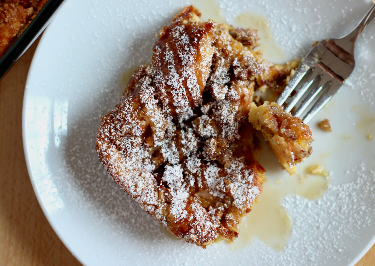 A piece of brioche french toast casserole on a small white plate with a fork removing a small piece of it.