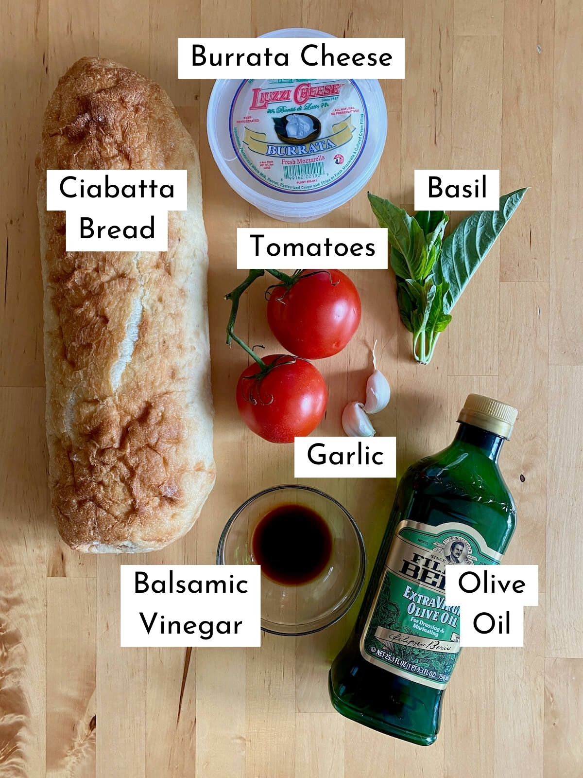 The ingredients to make burrata bruschetta. Each ingredient is labeled with text. They include ciabatta bread, burrata cheese, tomatoes, basil, garlic, balsamic vinegar, and olive oil.