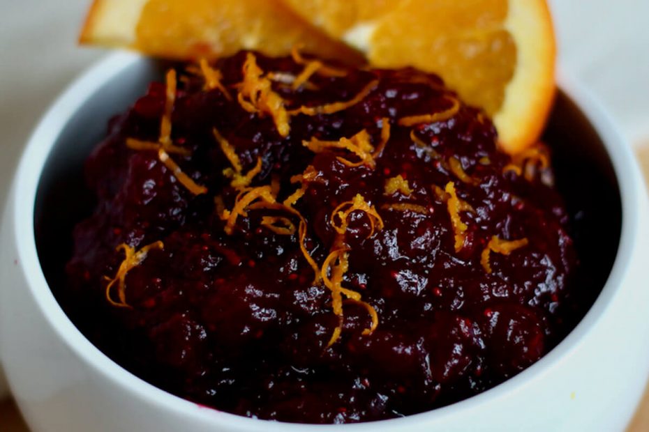 A small white bowl filled with cranberry sauce with orange juice. The cranberry sauce is garnished with fresh orange zest and two slices of orange.