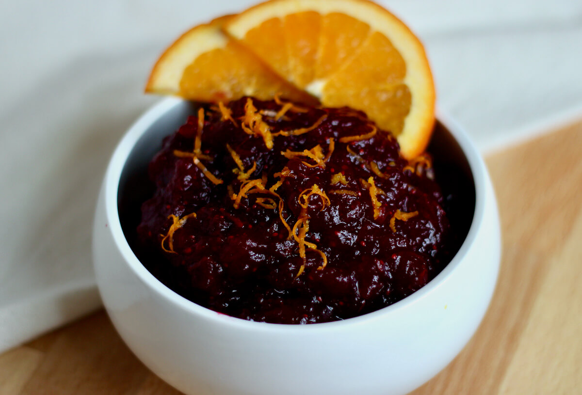A small white bowl filled with cranberry sauce with orange juice. The cranberry sauce is garnished with fresh orange zest and two slices of orange.