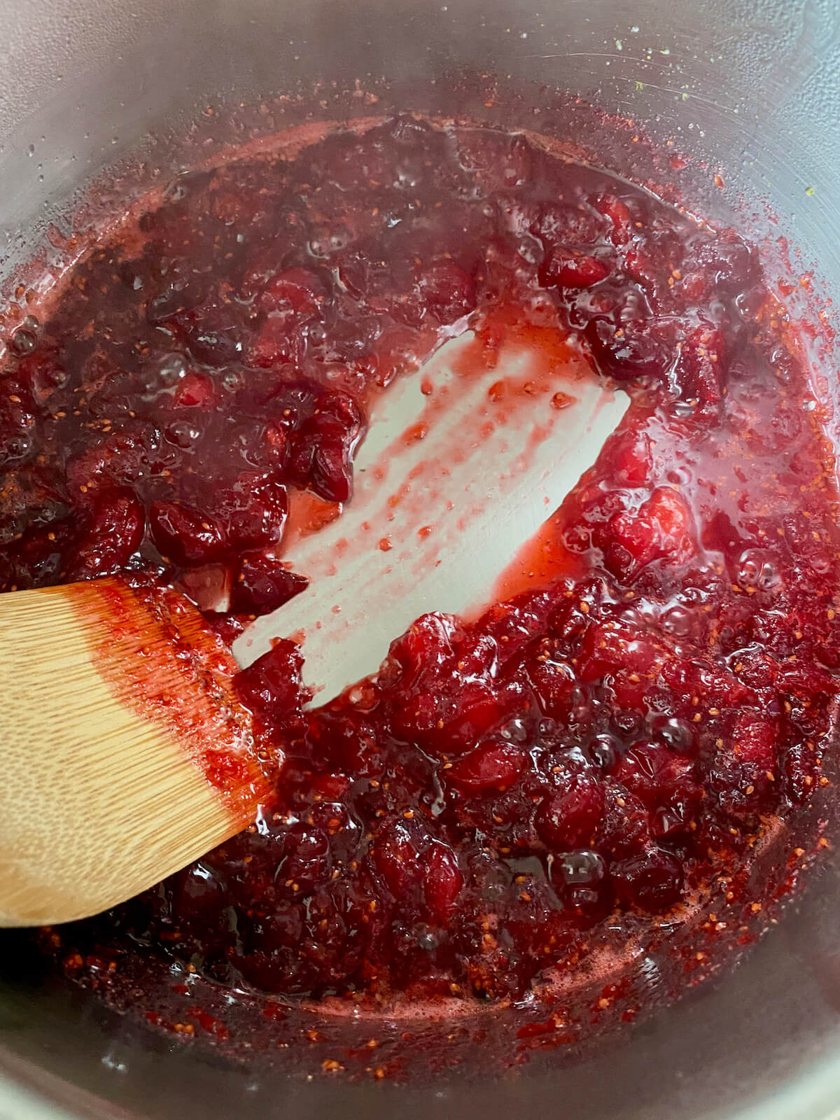 Homemade orange cranberry sauce thickened in a saucepan. There is a wooden spoon running through the sauce, revealing the bottom of the pot.