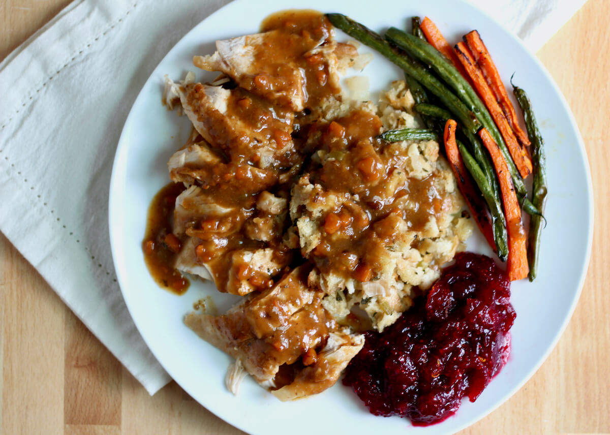 A large white plate filled with dutch oven turkey breast and gravy, stuffing, cranberry sauce, and roasted green beans and carrots.