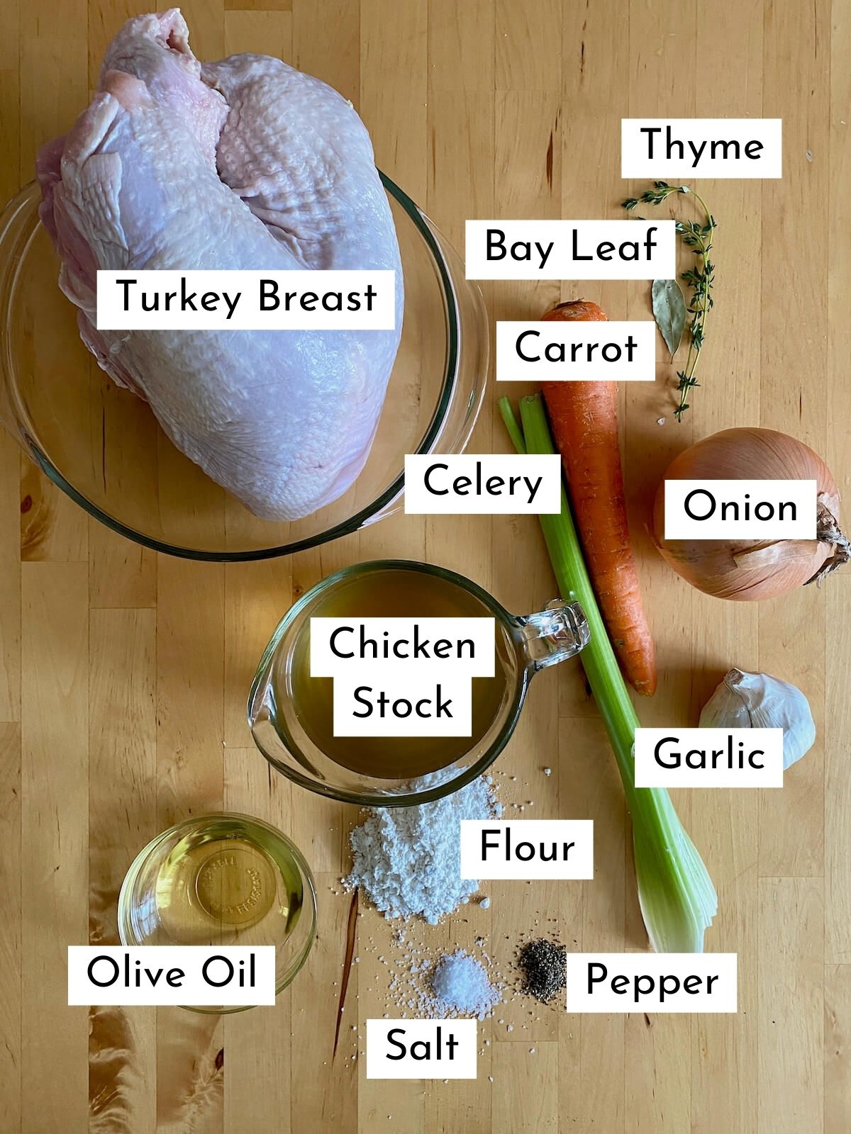 The ingredients to make dutch oven turkey breast on a butcher block countertop. Each ingredient is labeled with text stating what it is. They include turkey breast, bay leaf, thyme, carrot, celery, onion, chicken stock, garlic, olive oil, flour, salt, and pepper.