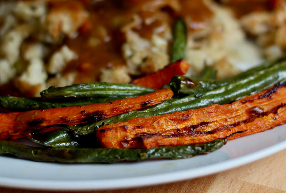 Roasted green beans and carrots on a large white plate. Out of focus in the background is stuffing and gravy.