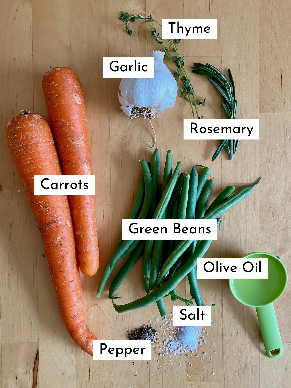 The ingredients to make roasted green beans and carrots on a butcher block countertop. Each ingredient is labeled with text stating what it is. They include carrots, green beans, garlic, thyme, rosemary, olive oil, salt, and pepper.