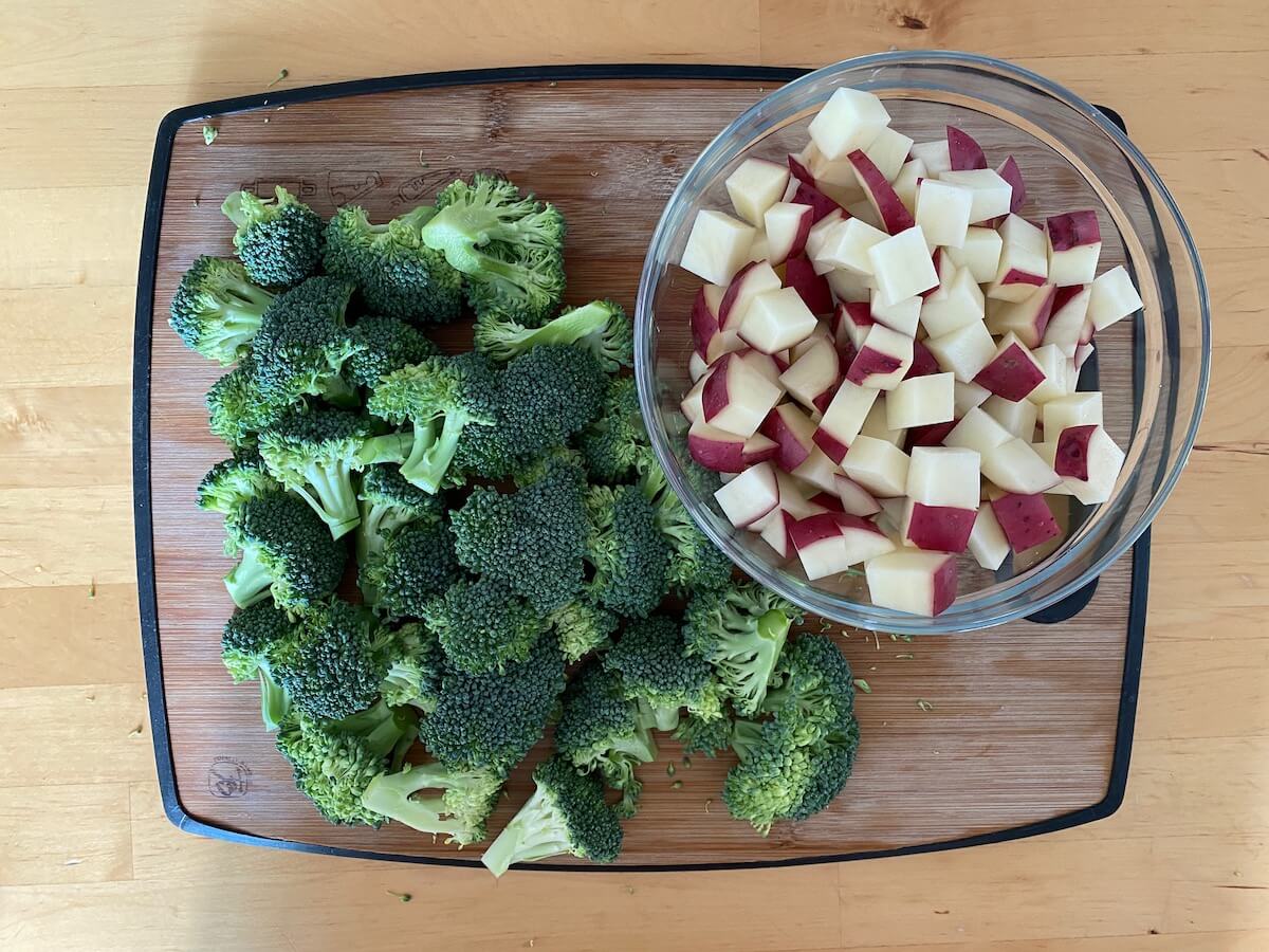 Broccoli florets and cubed potatoes on a cutting board. The potatoes are in a clear glass bowl, separate from the broccoli.