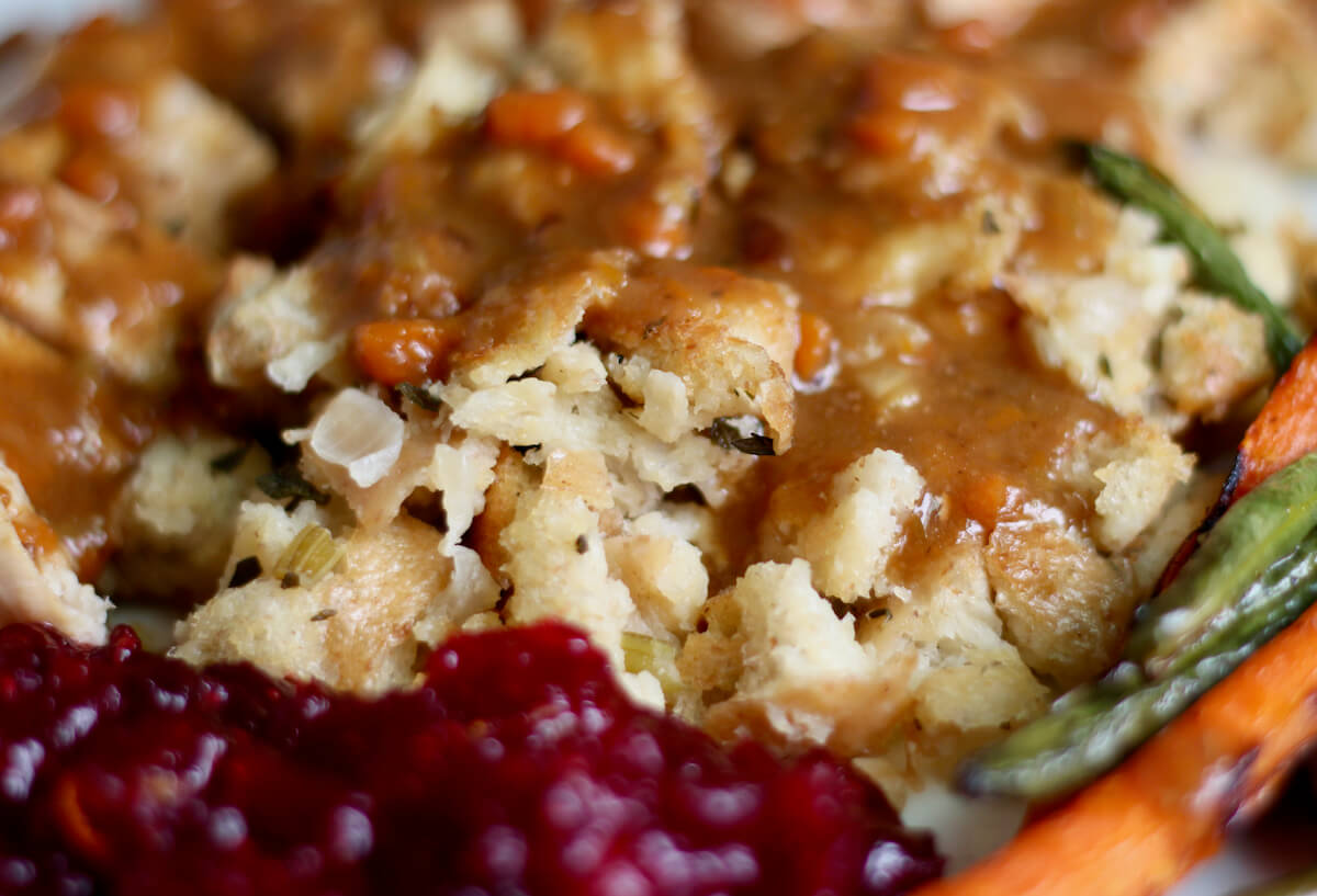 Sourdough stuffing topped with gravy next to cranberry sauce and vegetables on a large white plate.