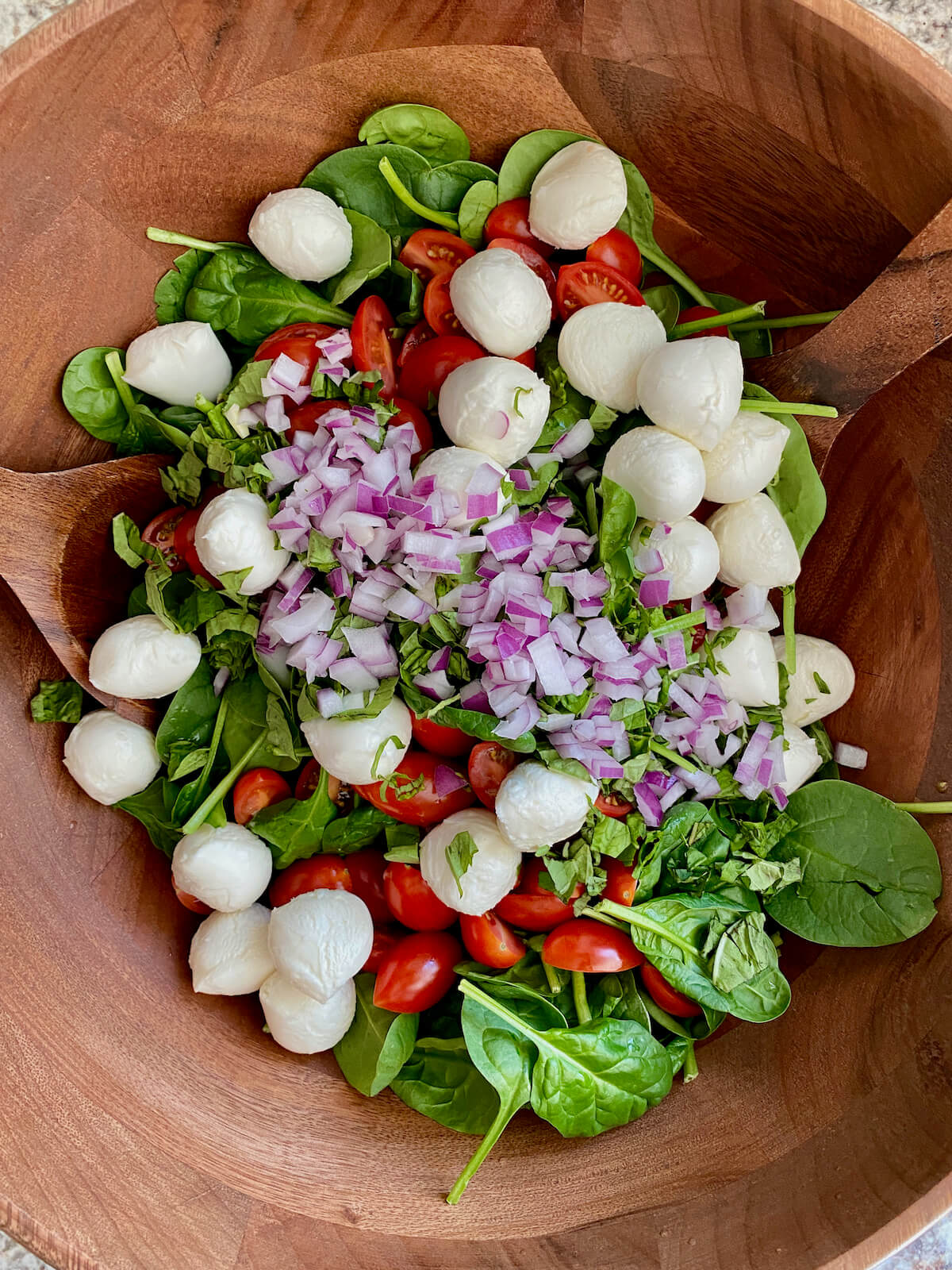 A large wooden salad bowl with baby spinach, cherry tomatoes, mozzarella cheese, basil, and diced red onion in it.