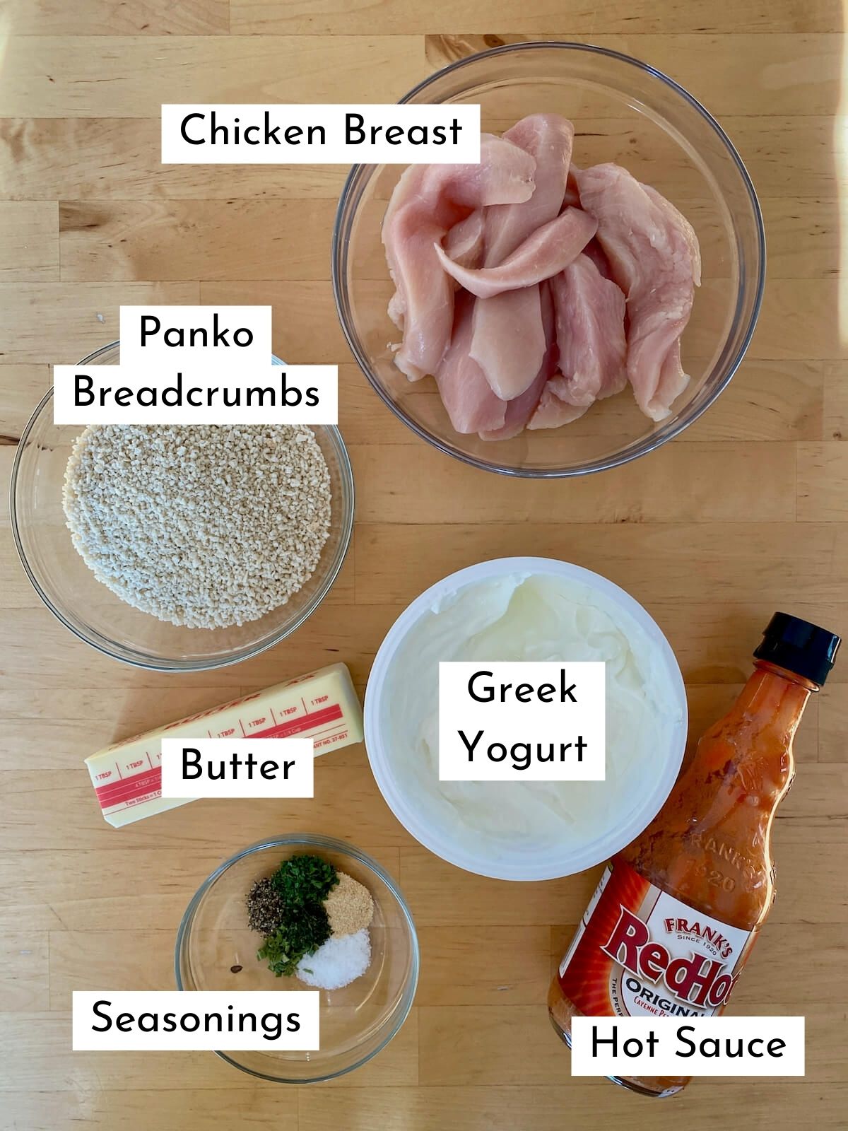 The ingredients to make air fryer buffalo chicken tenders. Each ingredient is labeled with text stating what it is. The ingredients include chicken breast, panko breadcrumbs, Greek yogurt, butter, seasonings, and hot sauce.
