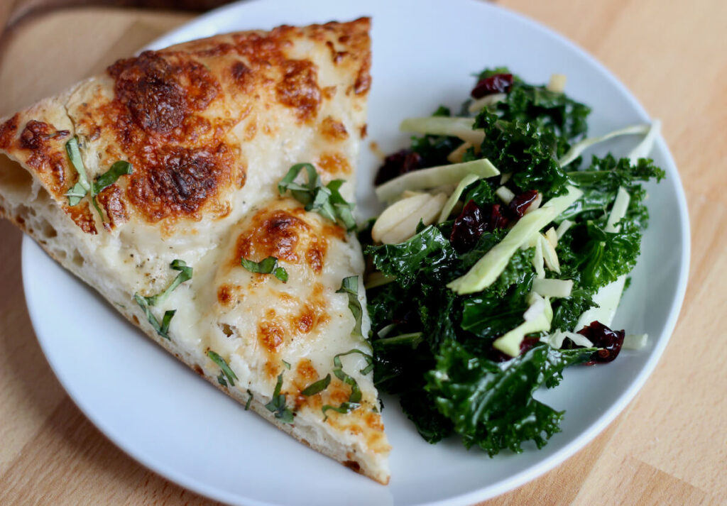 A slice of garlic parmesan pizza on a small white plate next to a portion of kale salad.
