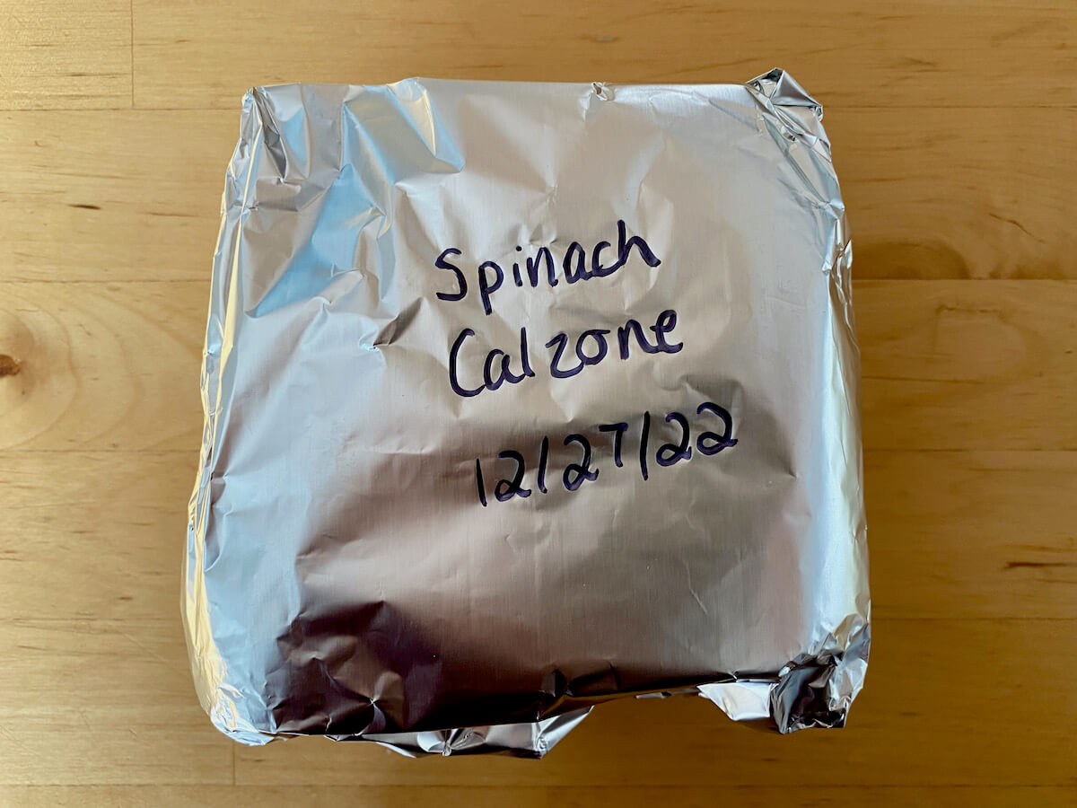 A piece of calzone wrapped in plastic wrap and aluminum foil. Written on the aluminum foil is "spinach calzone 12/27/22."