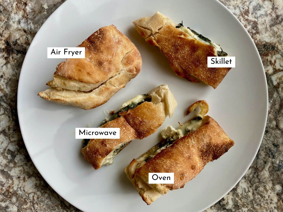 Four pieces of reheated calzone on a large white plate. There is text labeling the method used to reheat each piece of calzone. They include air fryer, skillet, oven, and microwave.