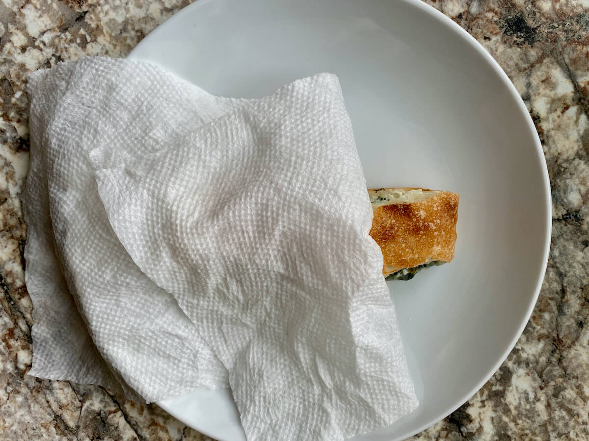 A piece of leftover calzone on a white plate with a damp paper towel covering most of it.