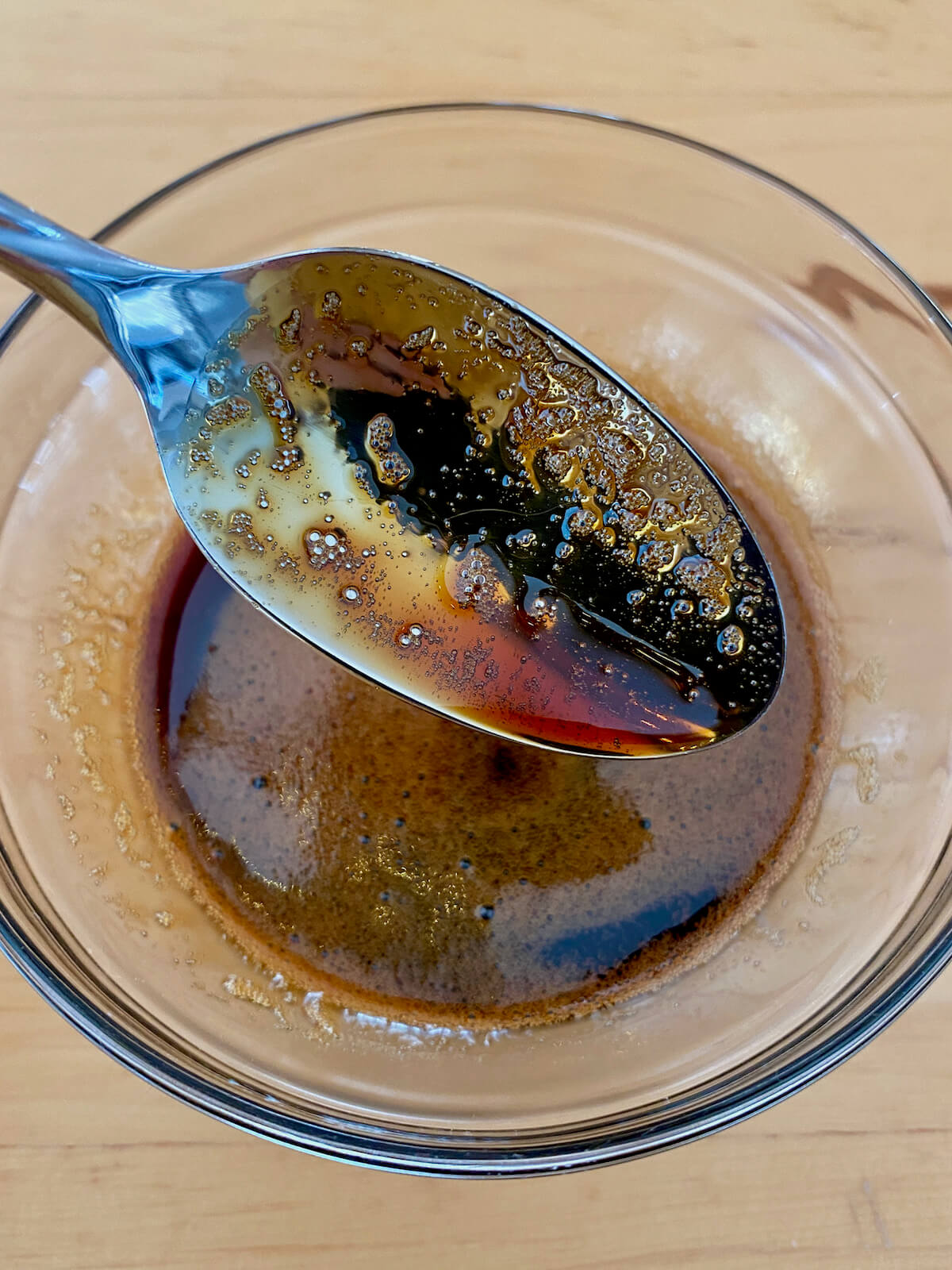A metal spoon holding up some maple balsamic glaze over a small glass bowl.