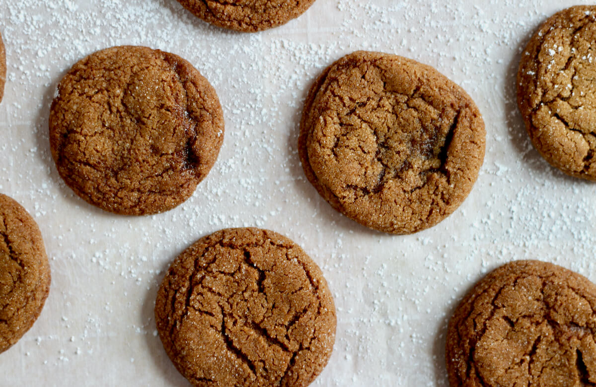 Molasses crinkle cookies evenly spaced on a sheet of parchment paper surrounded by powdered sugar.