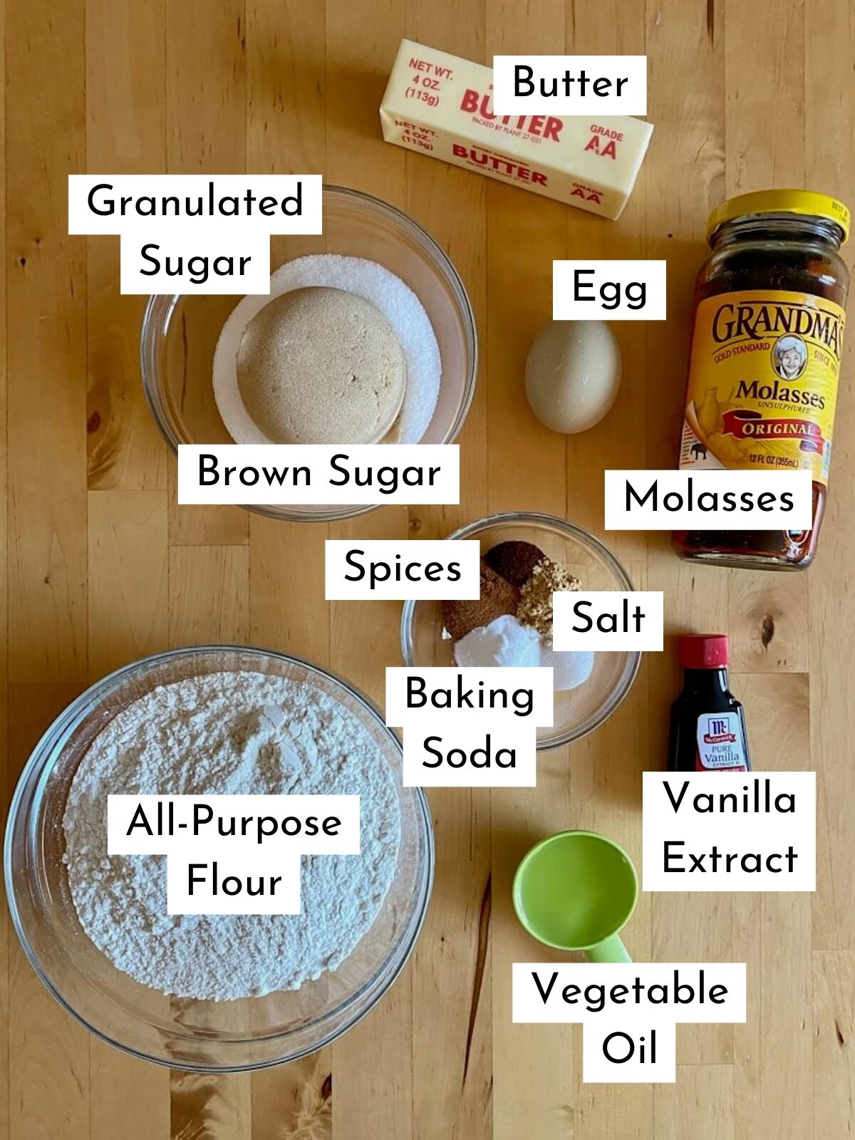 The ingredients to make molasses crinkle cookies. They're labeled with text stating what each ingredient is. They include butter, granulated sugar, brown sugar, egg, molasses, spices, salt, baking soda, all-purpose flour, vanilla extract, and vegetable oil.