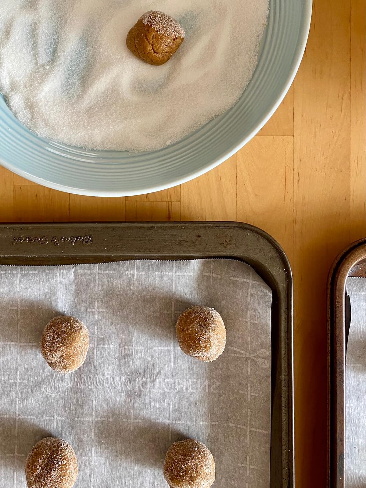 Molasses crinkle cookie dough balls being rolled and sugar and placed on a parchment-lined baking sheet.