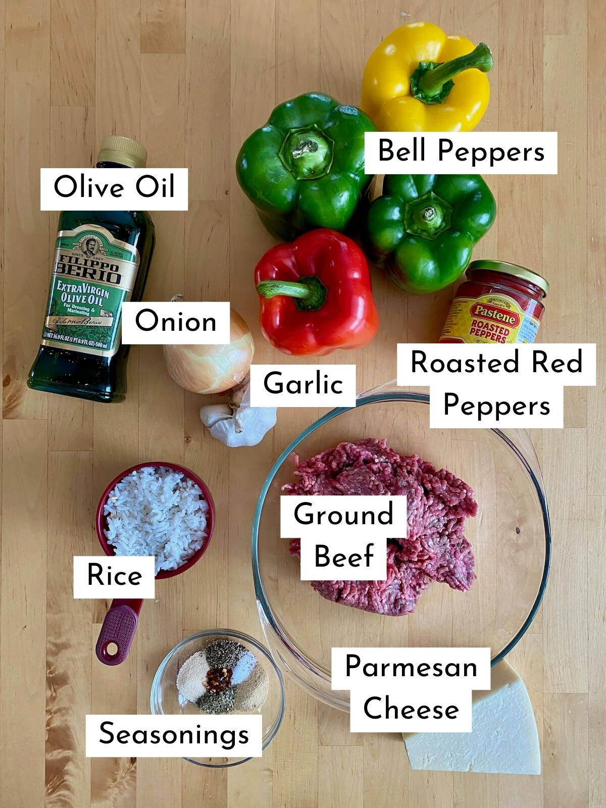 The ingredients to make stuffed peppers without tomato sauce. Each ingredient is labeled with text stating what it is. They include bell peppers, olive oil, onion, garlic, roasted red peppers, ground beef, rice, seasonings, and parmesan cheese.