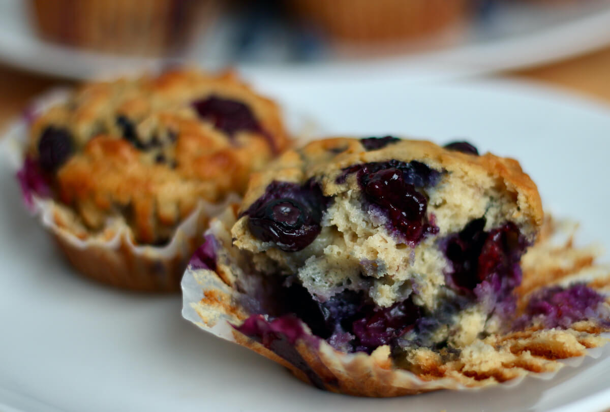 Two blueberry banana oatmeal muffins on a small white plate. One of the muffins is half eaten so you can see the inside texture.