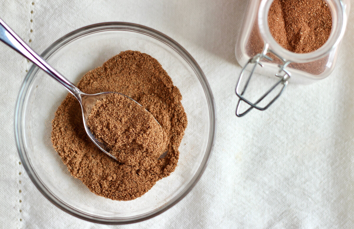 A small glass bowl filled with homemade chai spice blend. There is a spoon in the bowl of spices. To the right, there is a small spice jar filled with additional chai spice mix.