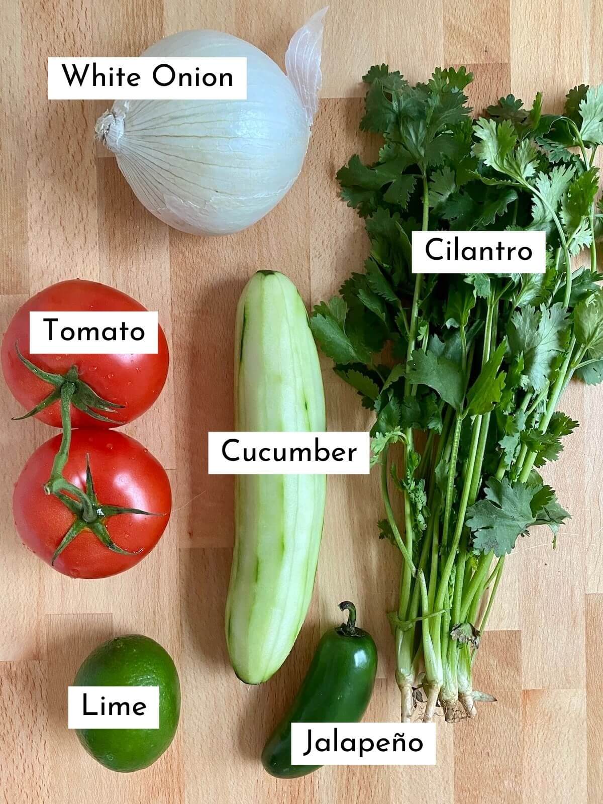 The ingredients to make cucumber pico de gallo. Each ingredient is labeled with text and includes white onion, tomato, cucumber, cilantro, lime, and jalapeño.
