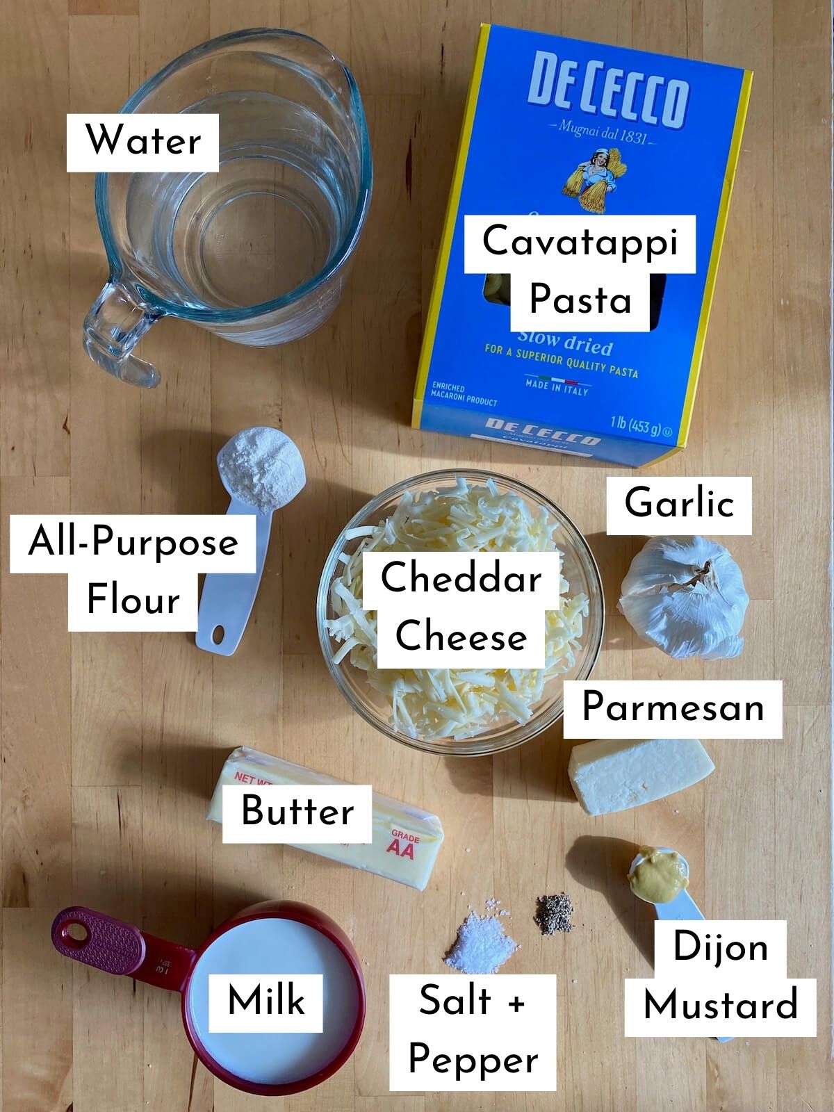 The ingredients to make dutch oven mac and cheese. The ingredients are labeled with text. They include water,  cavatappi pasta, all-purpose flour, cheddar cheese, garlic, butter, parmesan cheese, milk, salt, pepper, and dijon mustard.