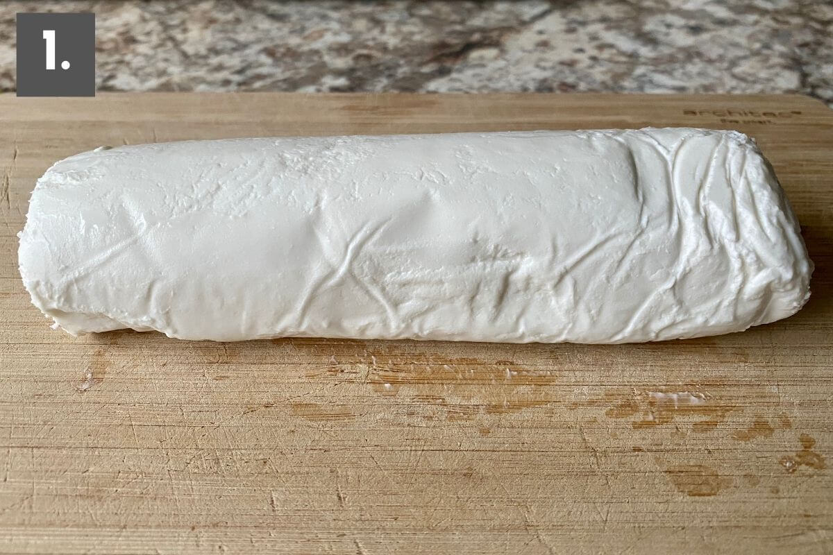 A solid log of plain goat cheese on a bamboo cutting board. There is a number one in the top left corner.