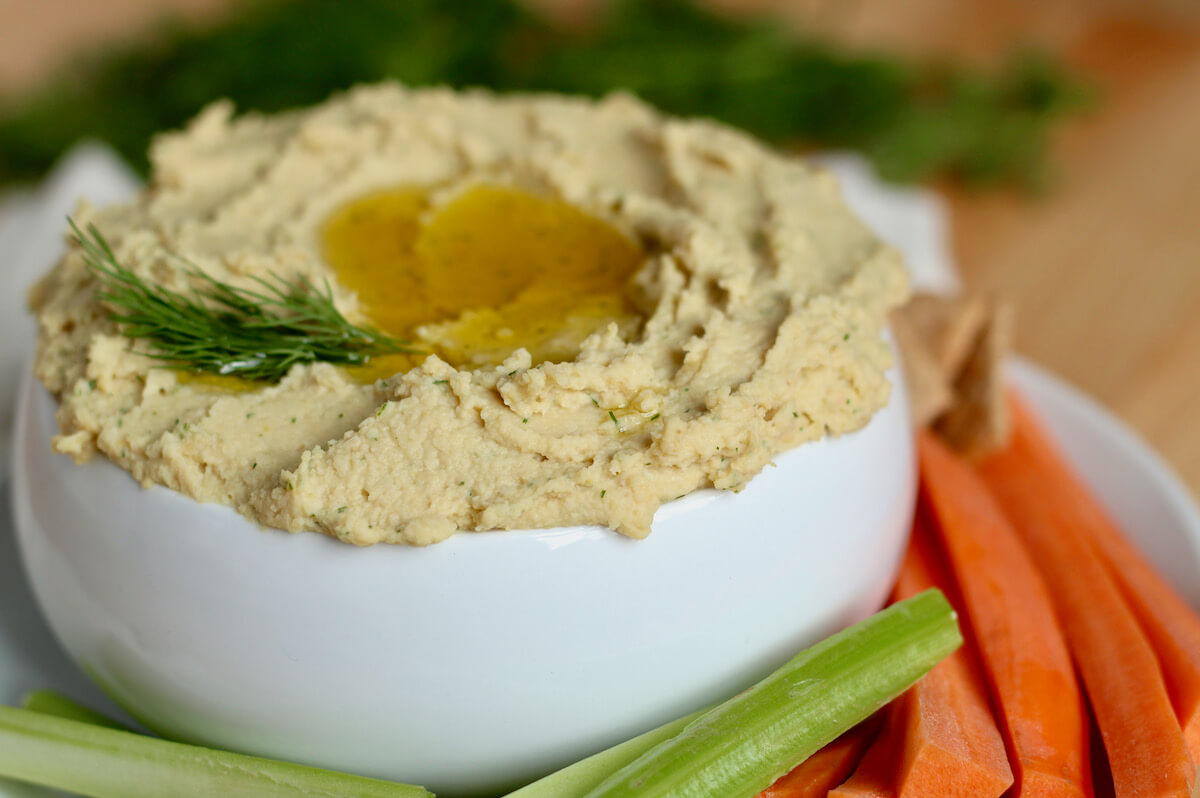 A bowl of lemon and dill hummus surrounded by vegetables and crackers.