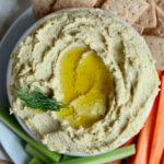 A bowl filled with lemon dill hummus topped with extra virgin olive oil and fresh dill. The bowl is surrounded by carrots, celery, and wheat crackers.