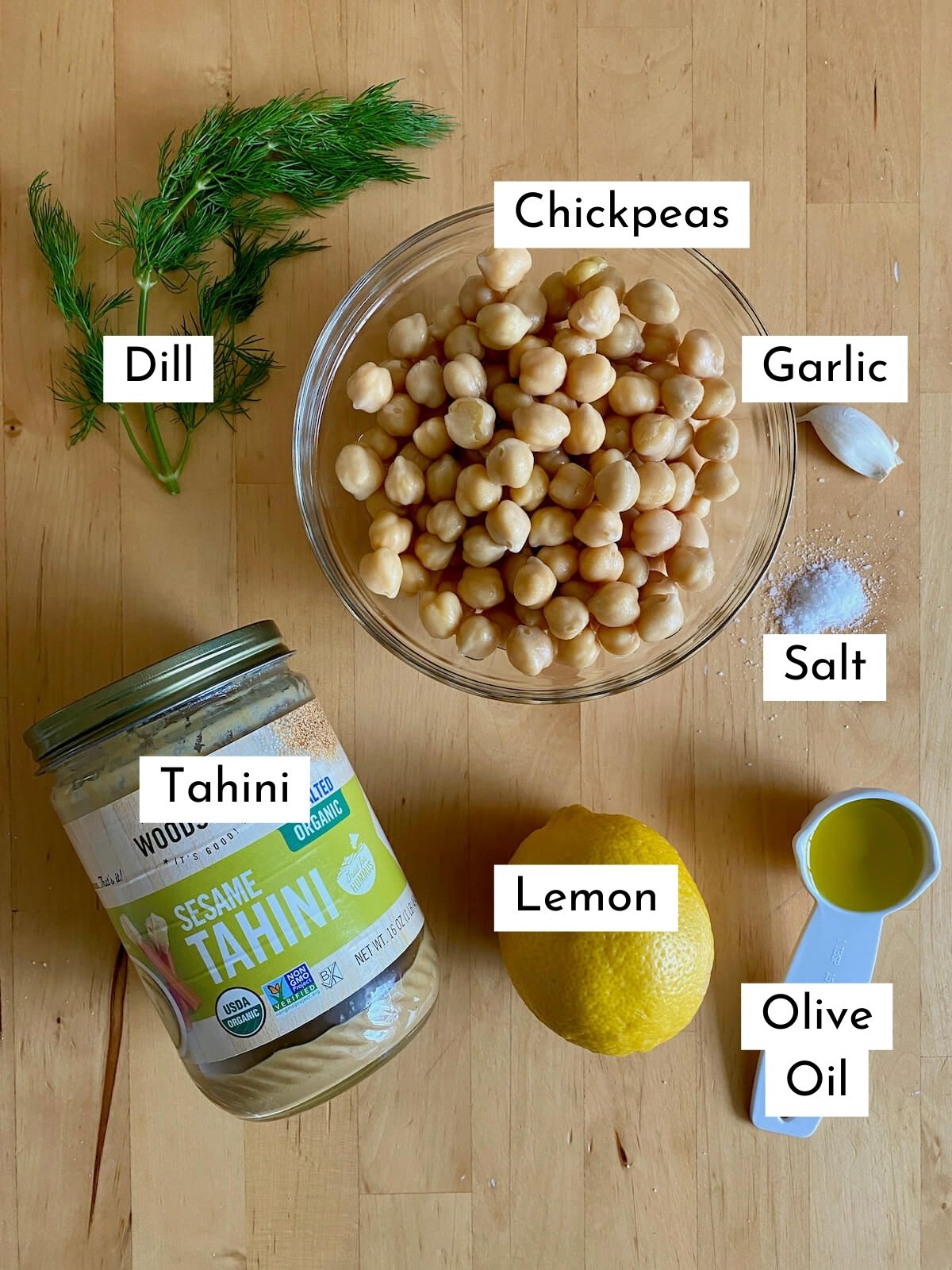 The ingredients to make lemon dill hummus. Each ingredient is labeled with text describing what it is. They include dill, chickpeas, garlic, tahini, salt, lemon, and olive oil.