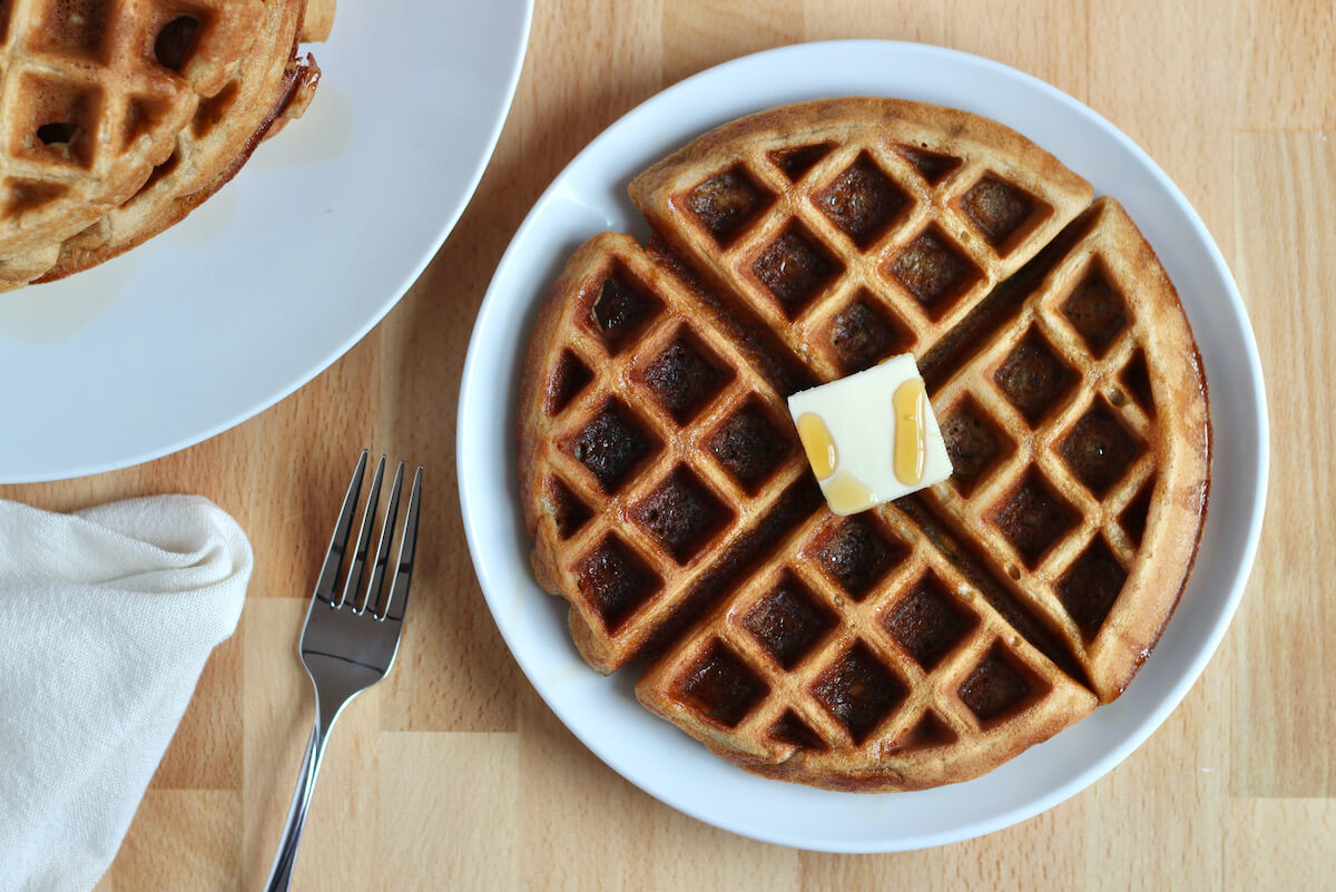 An oat flour waffle on a small white plate. The waffles is garnished with butter and maple syrup.