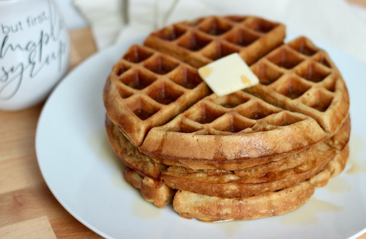 A stack of oat flour waffles with butter and maple syrup on a large white plate. There is a jar of maple syrup in the background.