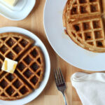 An oat flour waffle with butter and maple syrup on a small white plate. To the left of the plate is a stack of the remaining oat flour waffles.