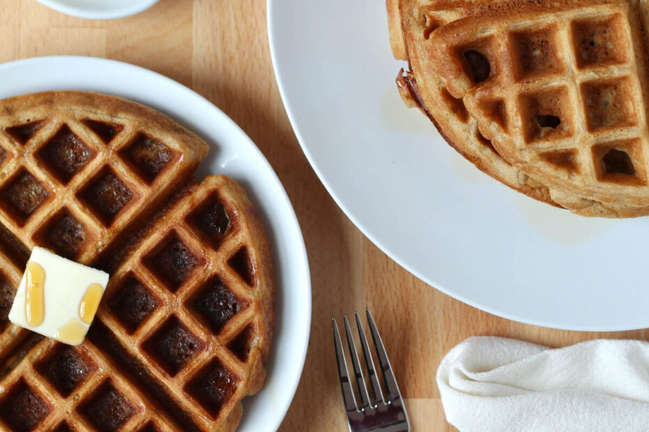 An oat flour waffle with butter and maple syrup on a small white plate. To the left of the plate is a stack of the remaining oat flour waffles.