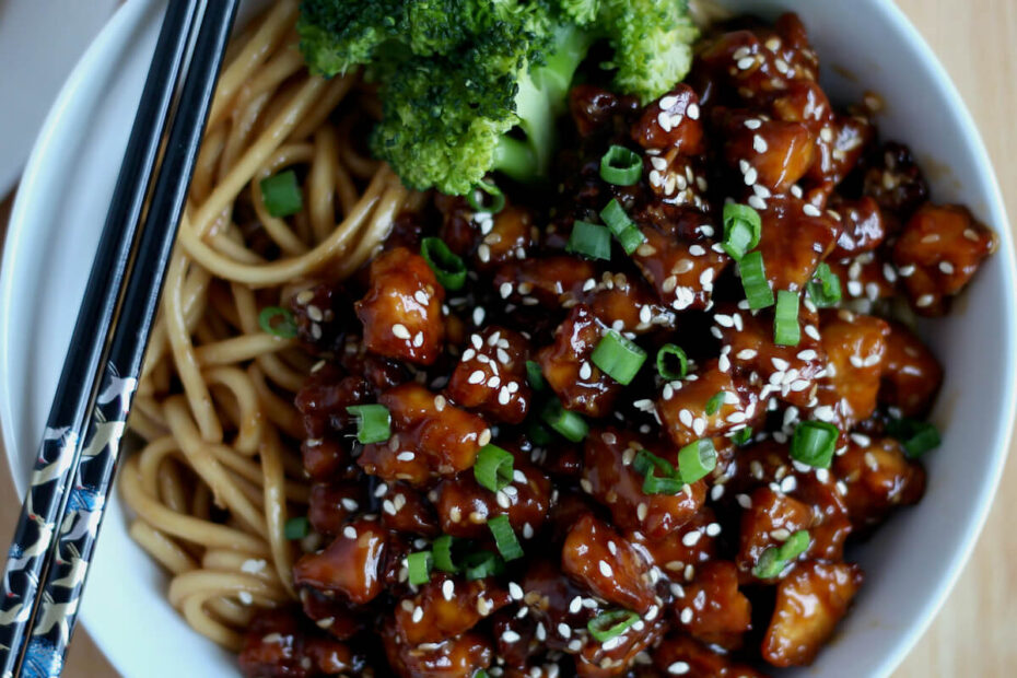 A bowl of sesame garlic tofu served with udon noodles and steamed broccoli. There is a pair of black chopsticks resting on the bowl.