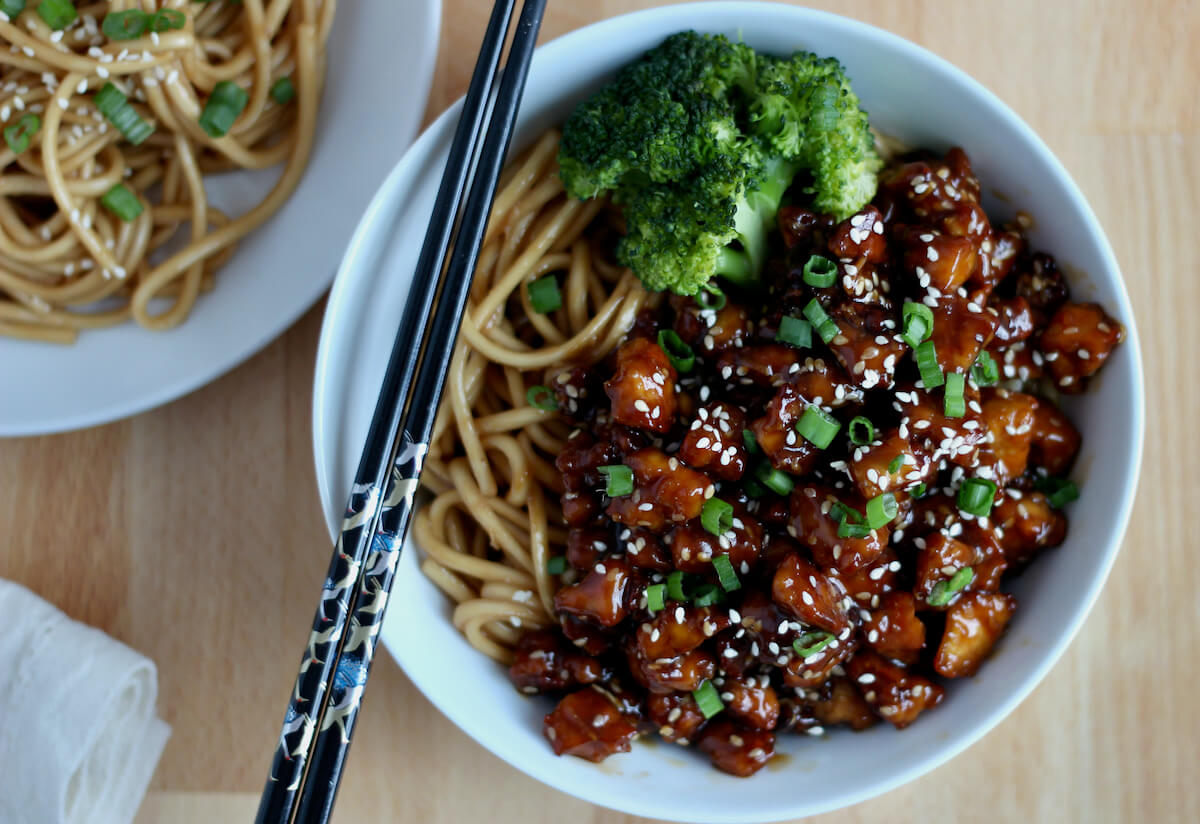 A bowl of sesame garlic tofu served with udon noodles and steamed broccoli. There is a pair of black chopsticks resting on the bowl. Out of focus to the left is an additional plate of noodles.