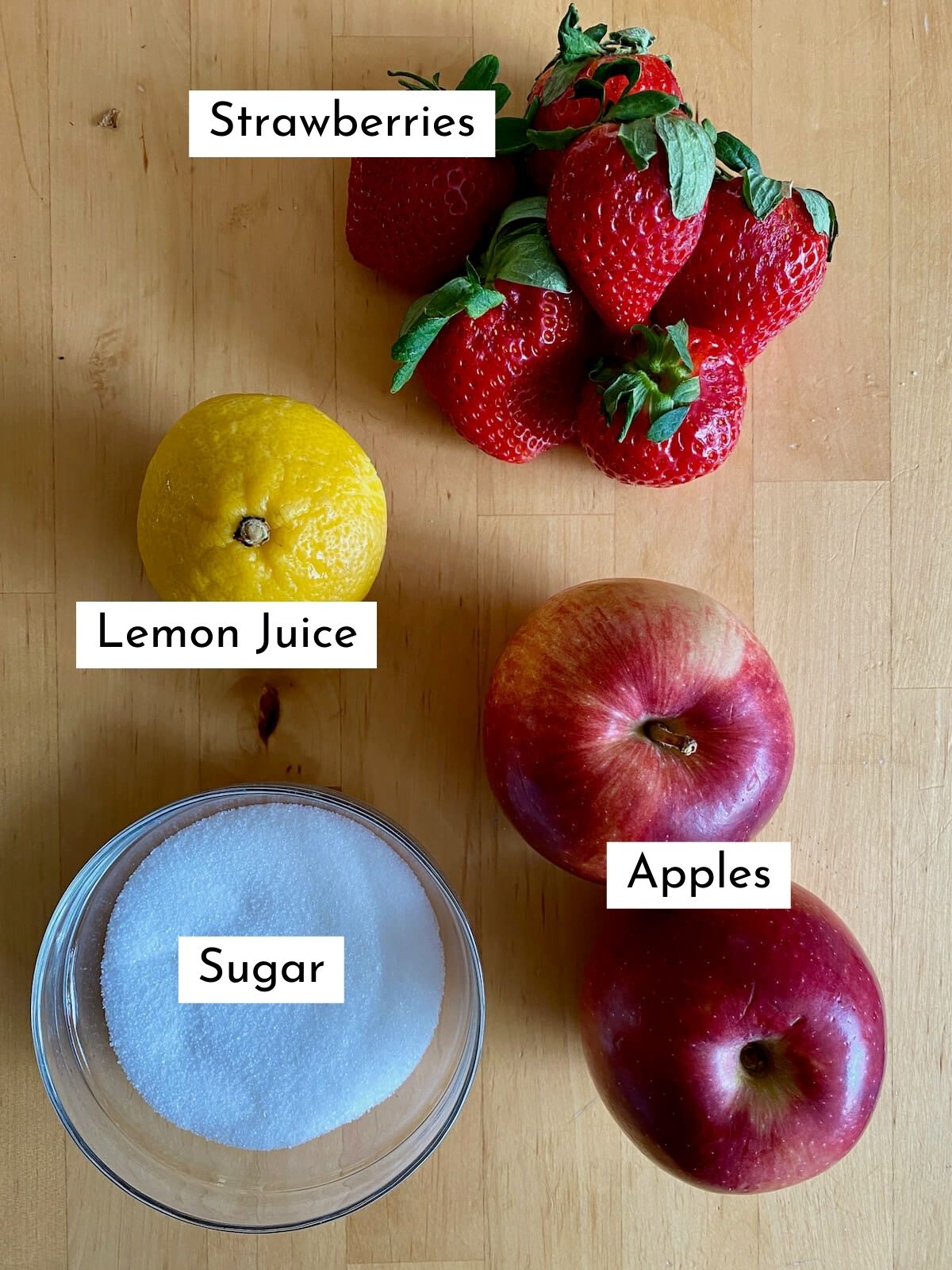 The ingredients to make strawberry apple jam on a butcher block countertop. Each ingredient is labeled with text describing what it is. They include strawberries, lemon juice, sugar, and apples.
