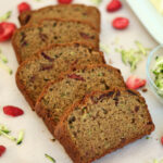 A sliced loaf of strawberry zucchini bread on a piece of parchment paper. Out of focus in the background is a small bowl of shredded zucchini, a butter dish, and a few pieces of freeze-dried strawberries.