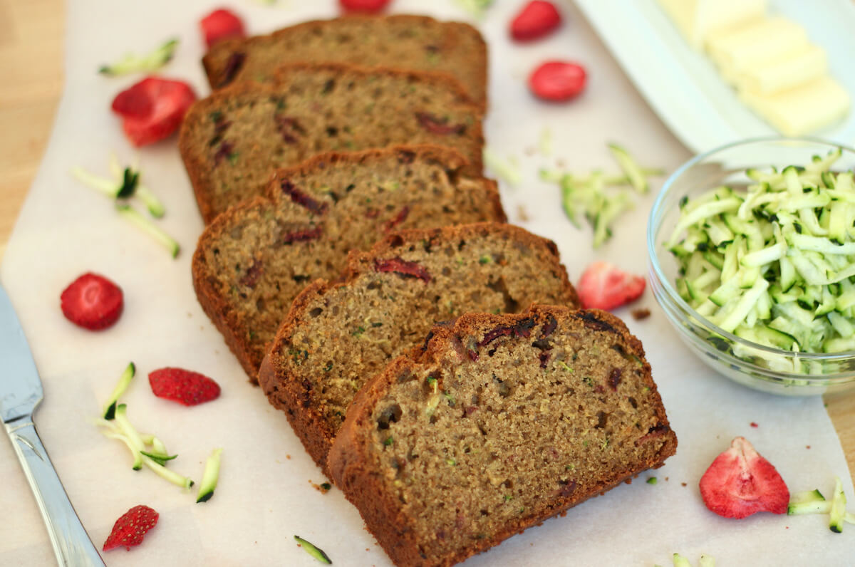 A sliced loaf of strawberry zucchini bread on a piece of parchment paper. Out of focus in the background is a small bowl of shredded zucchini, a butter dish, and a few pieces of freeze-dried strawberries.