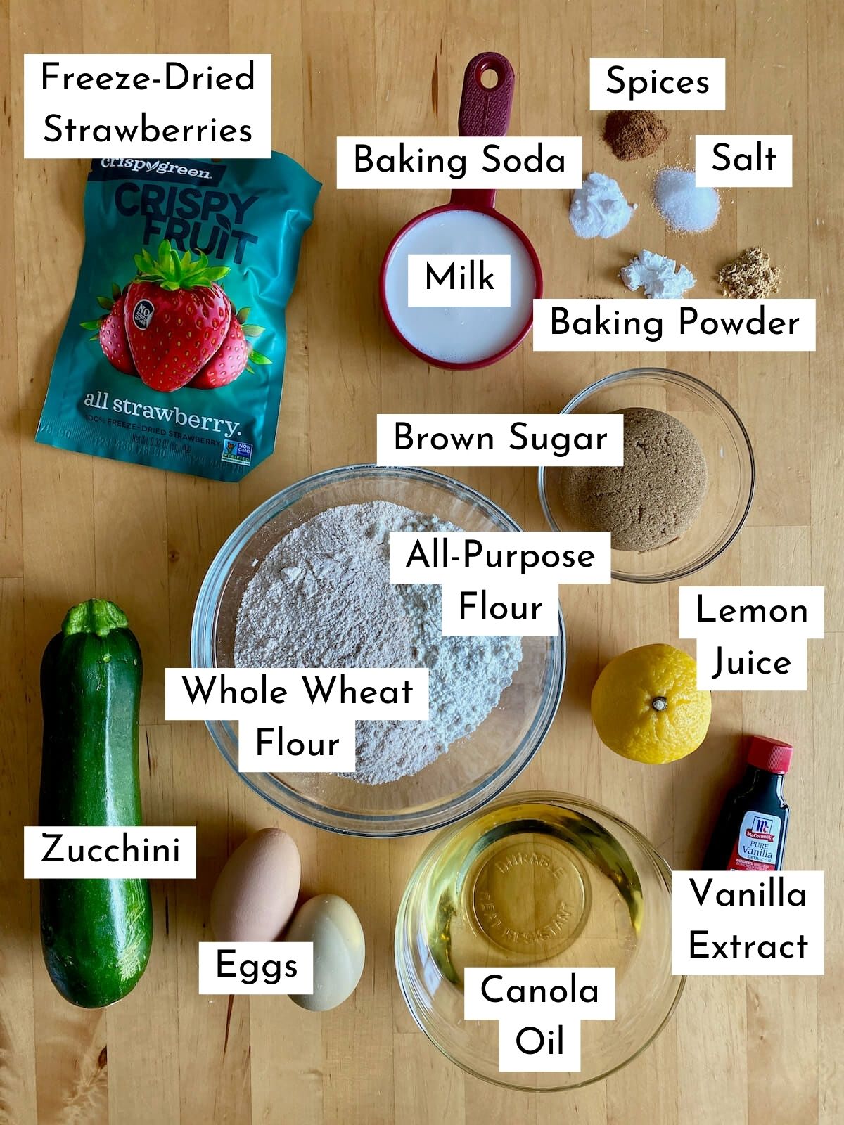 The ingredients to make strawberry zucchini bread on a butcher block counter. Each ingredient is labeled with text describing what it is. They include freeze-dried strawberries, baking soda, slices, salt, milk, baking powder, brown sugar, all-purpose flour, whole wheat flour, zucchini, lemon juice, eggs, canola oil, and vanilla extract.