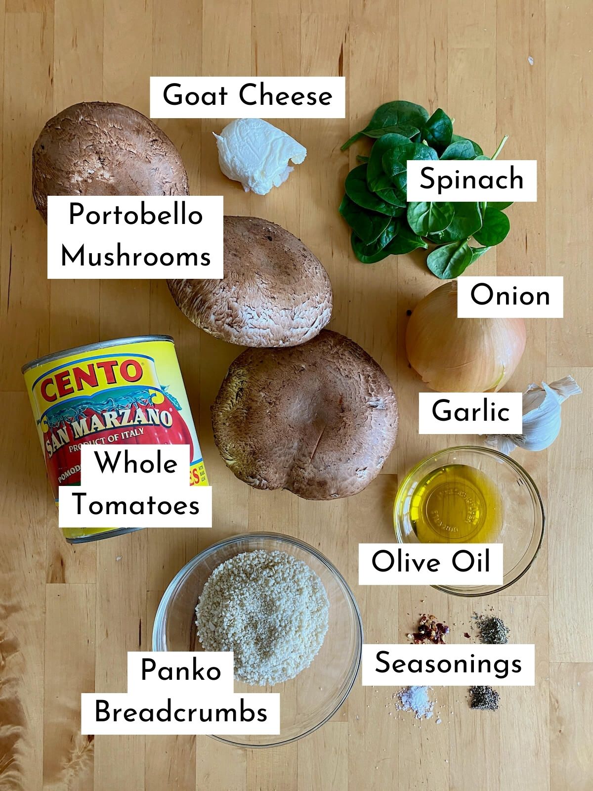 The ingredients to make stuffed portobello mushrooms with text labeling each ingredient. Ingredients include portobello mushrooms, goat cheese, spinach, whole tomatoes, onion, garlic, olive oil, panko breadcrumbs, and seasonings.
