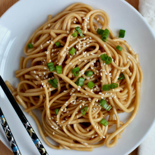 A small white plate with a pile of teriyaki udon noodles. The noodles are garnished with sesame seeds and chopped scallions. There is a pair of black chopsticks resting on the left side of the plate.