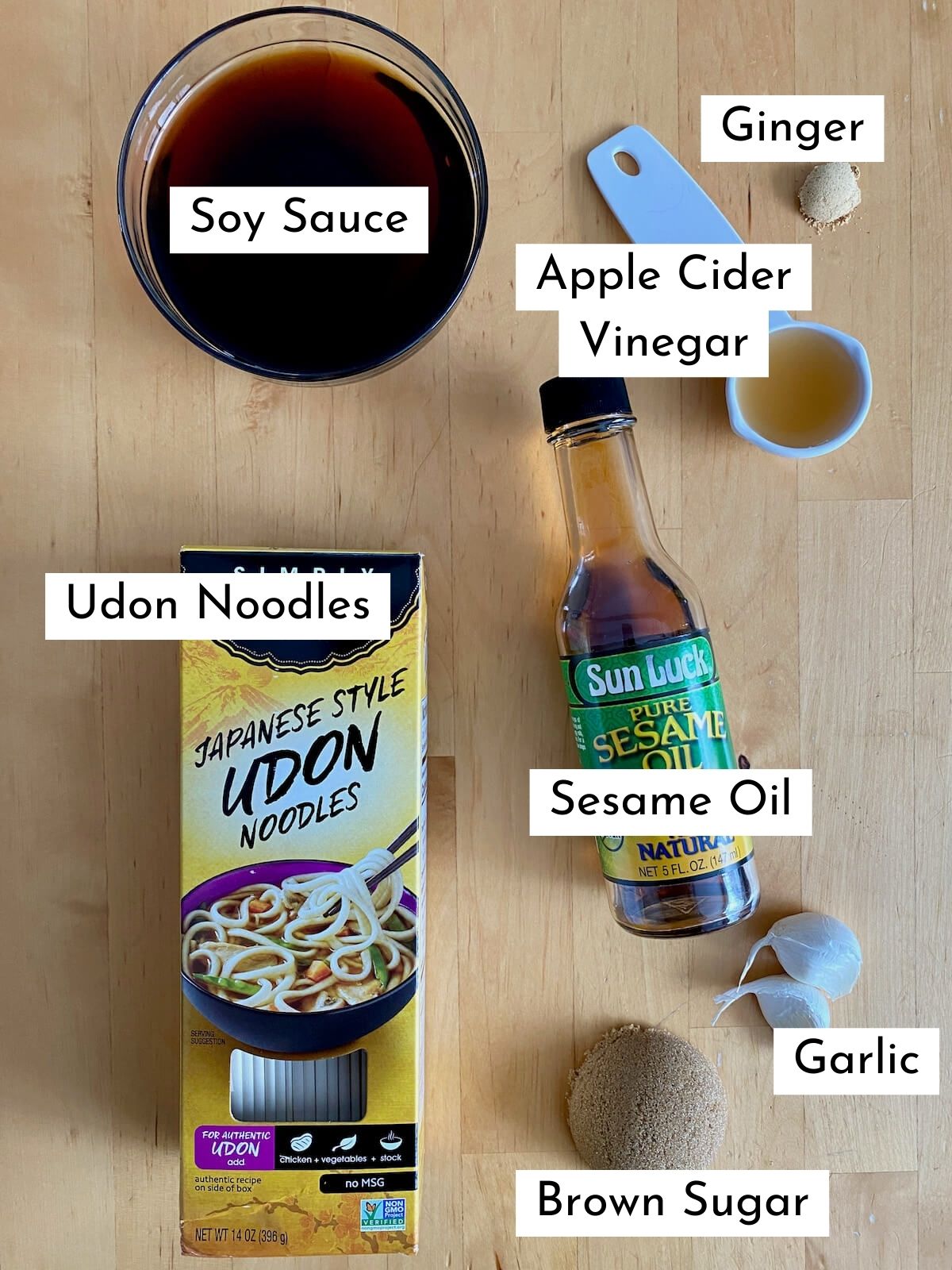 The ingredients to make teriyaki udon noodles. Each ingredient is labeled with text. They include soy sauce, apple cider vinegar, ginger, udon noodles, sesame oil, garlic, and brown sugar.
