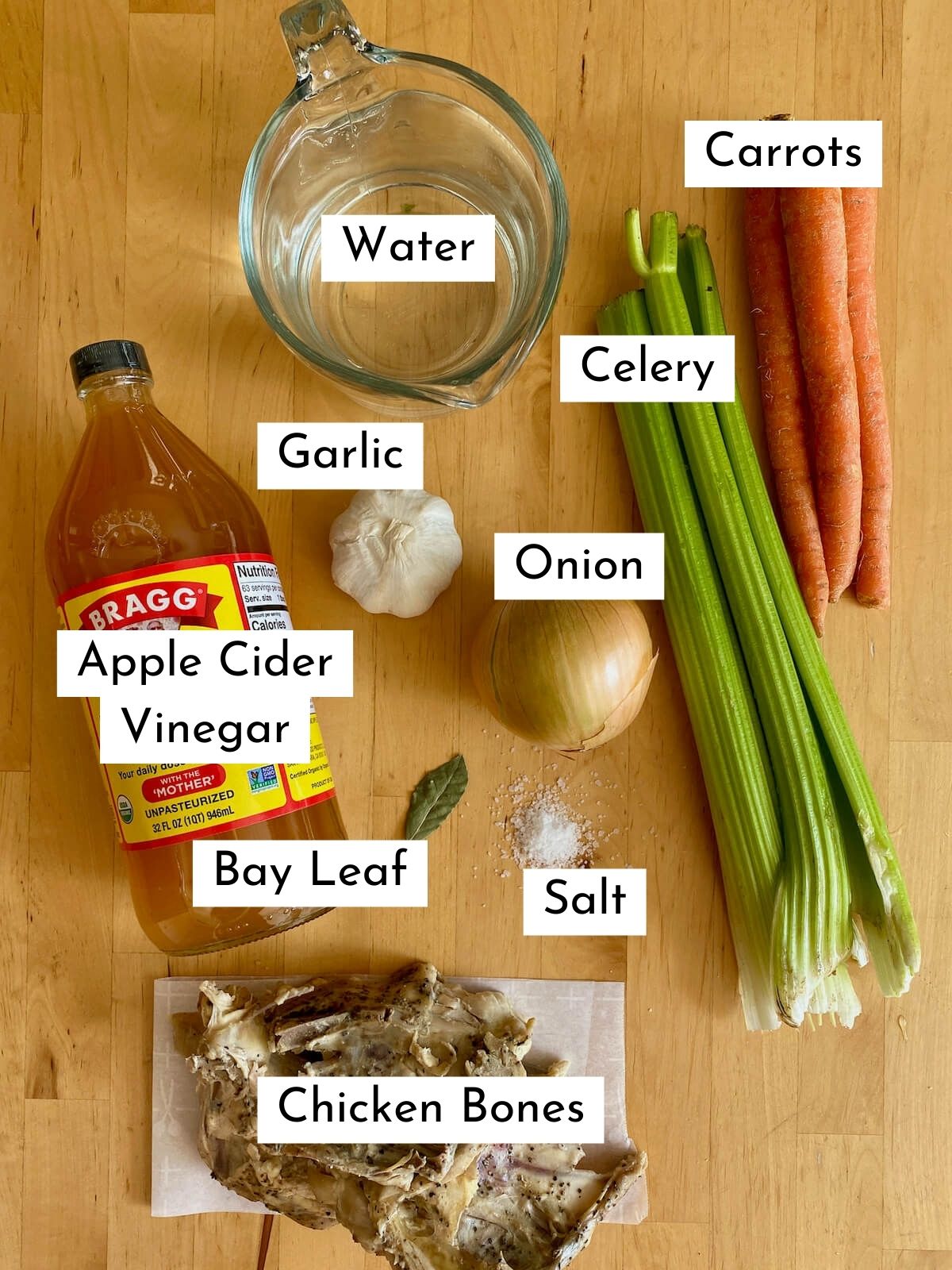 The ingredients to make chicken bone broth. Each ingredient is labeled with text. They include water, carrots, celery, garlic, onion, apple cider vinegar, bay leaf, salt, and chicken bones.