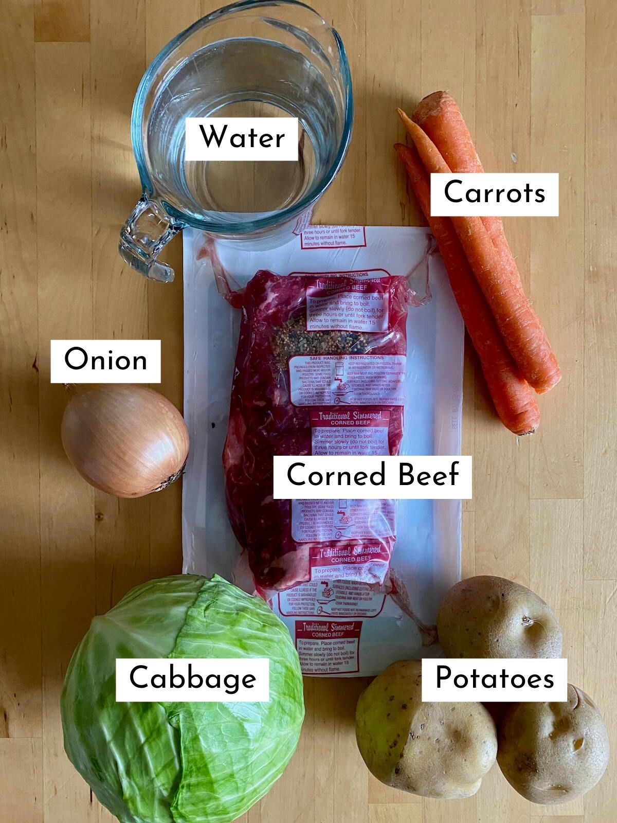 The ingredients to make Dutch oven corned beef and cabbage on a butcher block countertop. Each ingredient is labeled with text. Ingredients include corned beef, water, onion, carrots, cabbage, and potatoes.